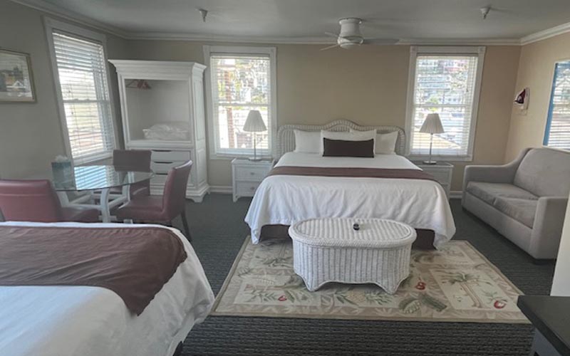 Two white beds in a bright hotel room, with a wicker coffee table on a floral rug, a beige sofa, and a glass dining table with red chairs nearby.